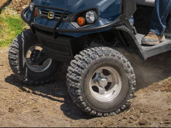 Close-up of a blue off-road utility vehicle with large knobby tires on a dirt ground, showing the front bumper, grille, and part of the driver's leg.